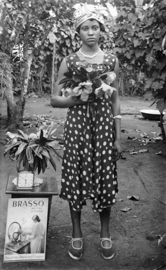 Mulher de pé com flores; relógio na mesa; "Brasso metal polish" [Dame Merry Oritsetimeyin Ehanire nee Cardigan (mãe de Osagie Ehanire)]