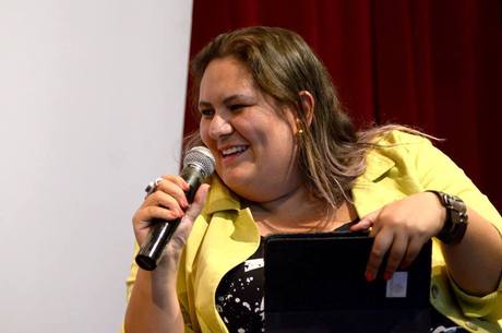 Jéssica Balbino- mulher branca de cabelo loiro que vai na altura dos ombros, vestindo um blazer amarelo- sentada sorrindo com um microfone na mão direita e uma pasta na mão esquerda. 