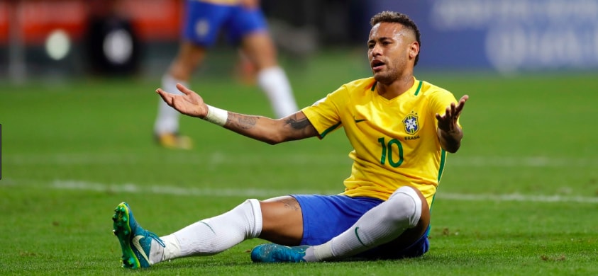 Neymar, homem negro, vestindo o uniforme da seleção brasileira, sentado no grama com as mão para cima (fazendo sinal de reclamação) durante uma partida de futebol 