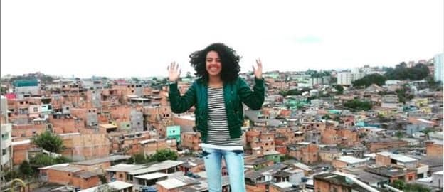 Veridiana Santana- mulher negra de cabelo cacheado, vestindo camiseta listrada, calça jeans e jaqueta verde- em pé sorrindo. Ao fundo uma comunidade 