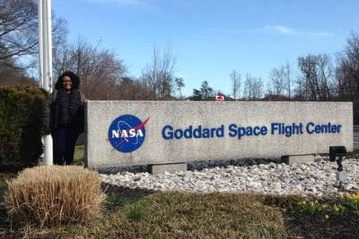  Carolyne Oliveira- mulher negra, de cabelo cacheado, usando roupas de frio- ao lado da placa do instituto da NASA  