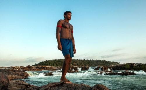 Giliard Juruna, homem indígena, sem camiseta e vestindo shorts azul escuro- em pé olhando para frente