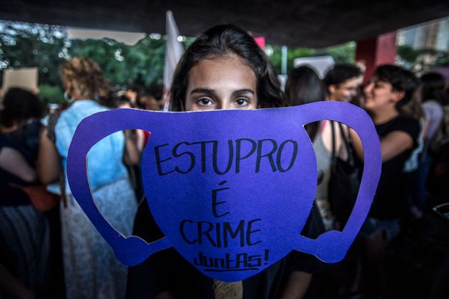 Todo de uma mulher com um cartaz na frente do rosto escrito "Estupro é crime, Juntas!"