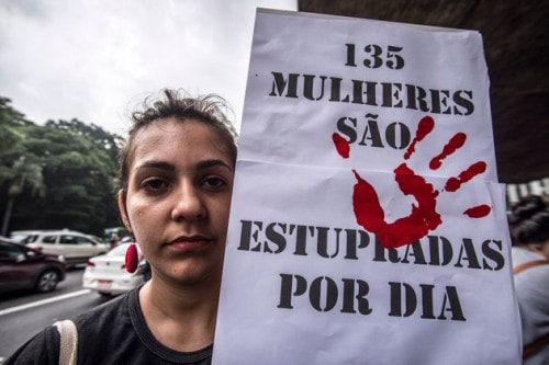 Uma mulher- branca, de cabelo preso- segurando um cartaz escrito "135 Mulheres são estupradas por dia"