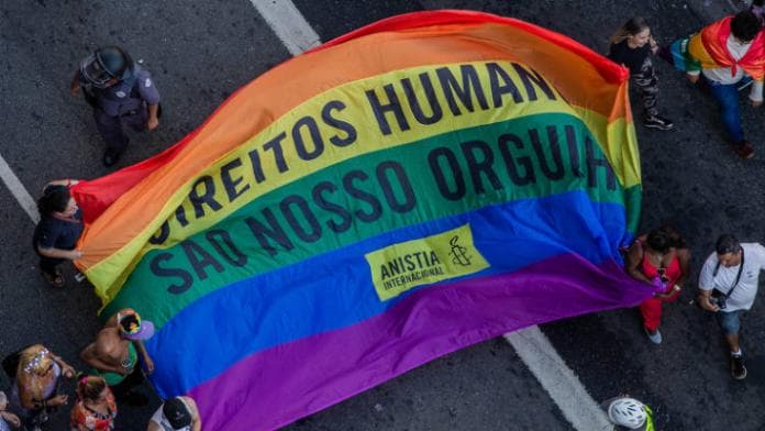 pessoas segurando uma bandeia LGBT escrito "Direitos Humanos são nosso Orgulho"