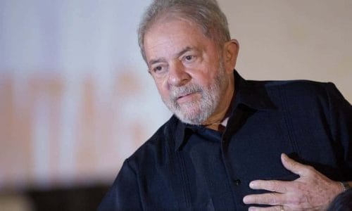 Luiz Inácio Lula da Silva- homem idoso branco, com barba e cabelos grisalhos, vestindo camisa social preta - em pé com a mãe esquerda sobre o peito 