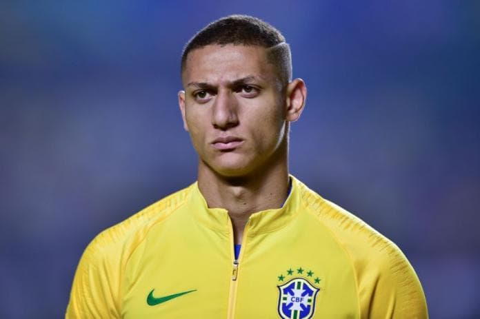 Richarlison- homem pardo vestindo o uniforme da seleção Brasileira- em pé olhando para frente 