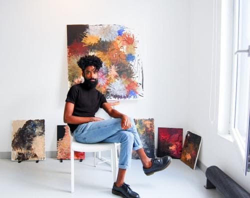 Elian Almeida- homem negro, barbudo, usando camiseta preta e calça jeans- sentado em uma cadeira cercado por seus trabalhos