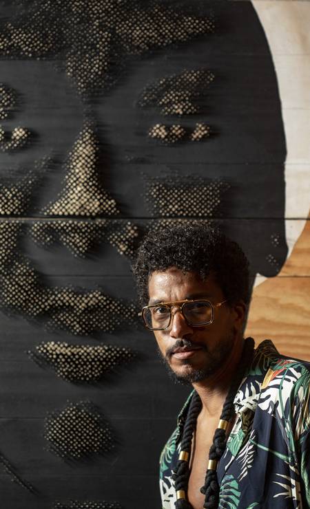 O artista Alexis Peskine - homem negro, usando óculos e camista colorida- em pé ao lado de uma pintura 