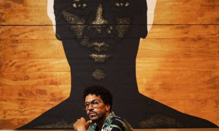 O artista Alexis Peskine - homem negro, usando óculos e camista colorida- agachado no chão 