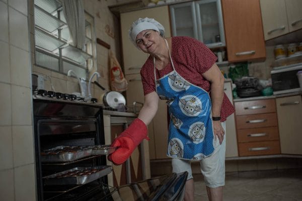 Rita Urquidi- mulher branca, usando luvas de cozinha, avental e touca - em pé tirando formas do forno