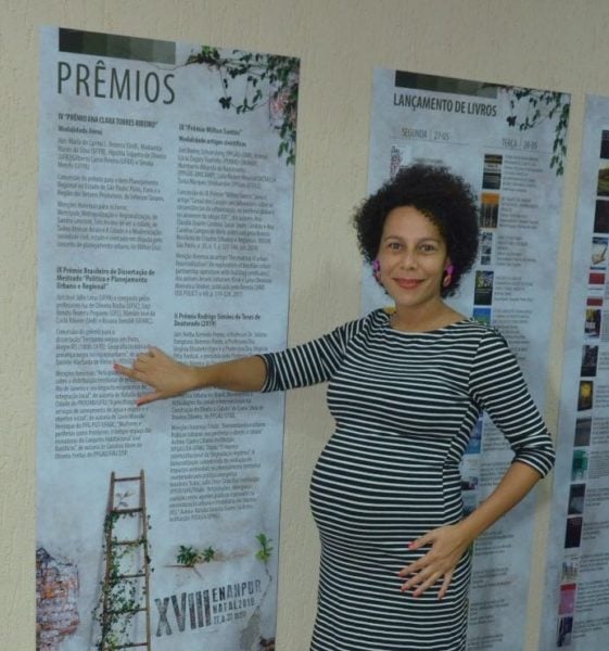  Daniele Vieira - mulher negra grávida, de cabelo curto cacheado, usando vestido listrado branco e preto - em pé, apontando para um cartaz na parede 