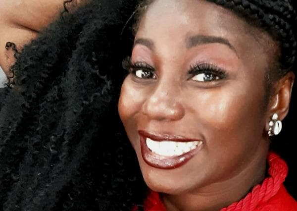 Carla Akotirane - mulher negra d cabelo cacheado, usando camiseta e batom vermelho-  sentada sorrindo  