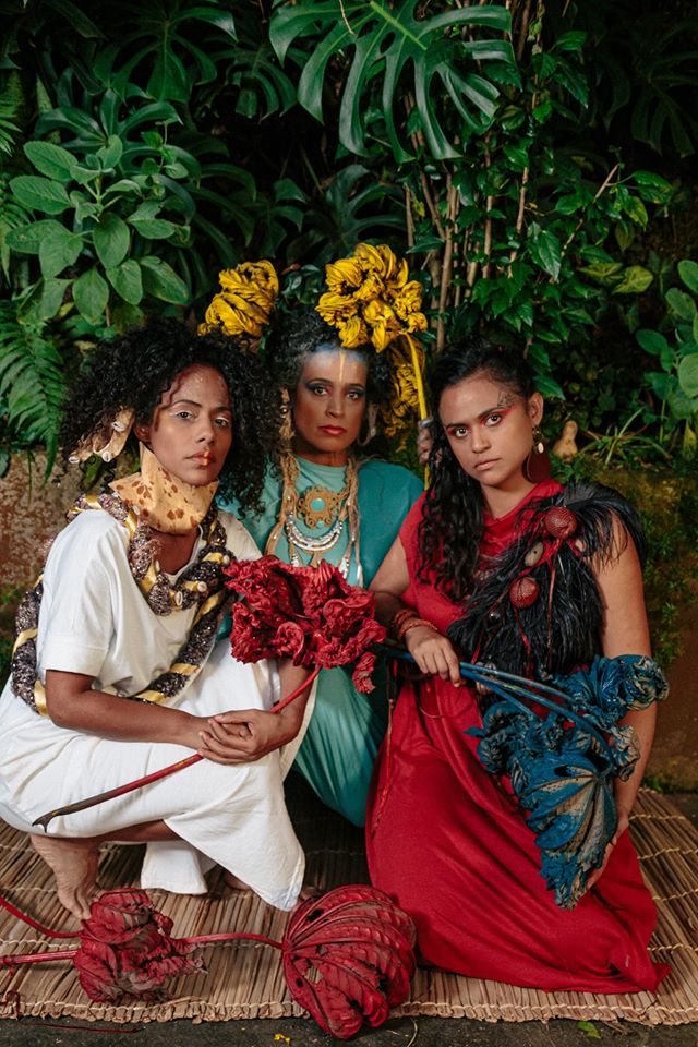 Trio musical Clarianas: Naruna Costa com as parceiras Martinha Soares e Naloana Lima - todas mulheres negras, vestindo roupas coloridas 