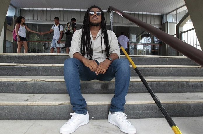 Miguel - homem negro de dreads, usando camiseta e calça jeans - sentado em uma escada 