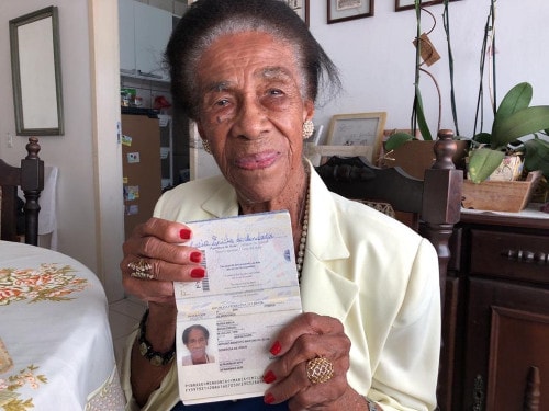 Maria Emília - mulher negra idosa, de cabelo grisalho, usando terninho branco- sentada segurando seu passaporte de 102 anos