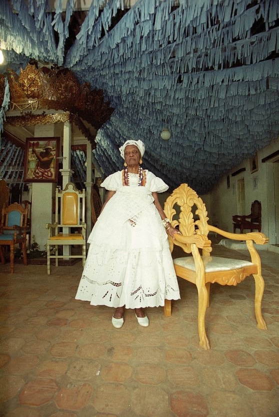 Mãe Tatá de Oxum- mulher negra idosa, vestino vestimentas brancas- em pé ao lado de uma cadeira 