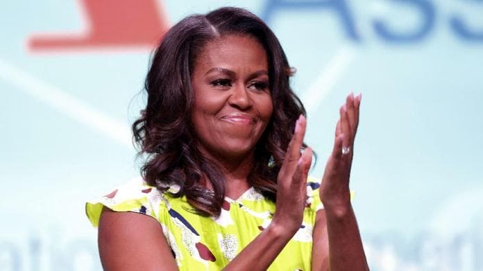 Michelle Obama- mulher negra, de cabelo liso, usando camiseta amarela com estampas coloridas- aplaudindo 