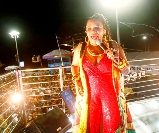 A cantora Margareth Menezes vestindo um luxuoso macacão vermelho em paetê e um max colete dourado com recortes assimétricos em preto e verde