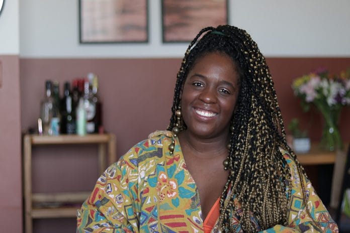 Ana Paula Xongani - mulher negra, usando tranças e camiseta com estampa colorida- sentada sorrindo 