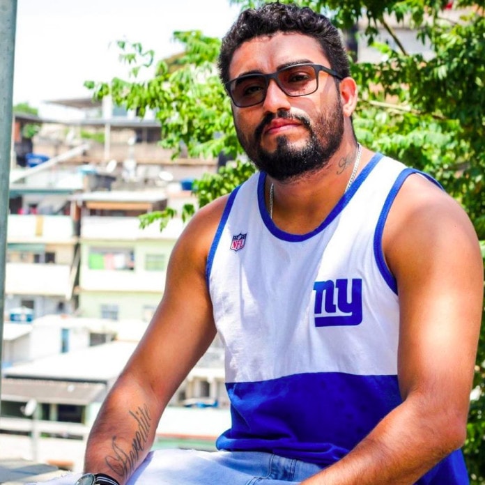 Raul Santiago- homem negro de barba e cabelo, usando óculos de sol e camiseta branca e azul- sentado olhando para frente 