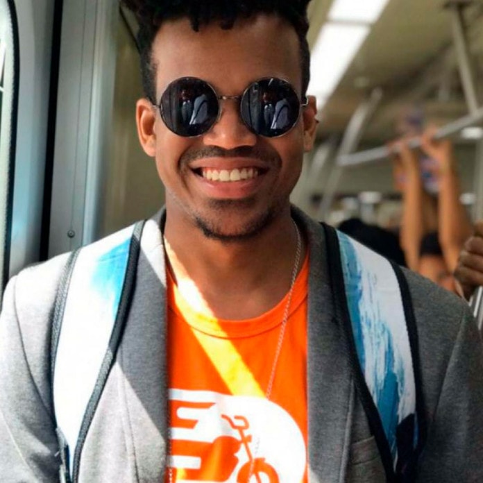 Iago Santos - homem negro de cabelo curto e pouca barba, usando óculos de sol e camiseta laranja- em pé sorrindo no metro