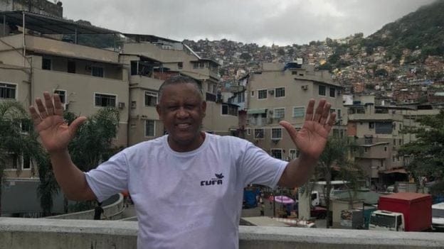 Celso Athayde - homem negro de pouco cabelo, vestindo camiseta branca- em pé sorrindo e acenando com as duas mãos 