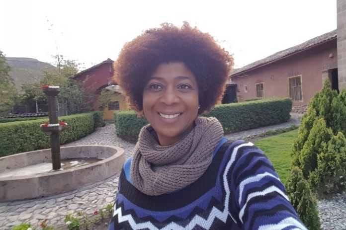 Selfie de Rosane de Souza- mulher negra, de cabelo crespo, usando blusa de frito com estampa, preto, azul e branco e um cachecol marrom- em pé sorrindo 