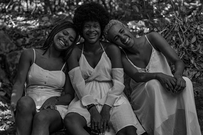 Foto em preto e branco de três mulheres negras com cabelos distintos- a primeira usa tranças, a segunda black e a terceira tem o cabelo bem curto - sentadas sorrindo 