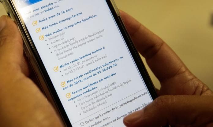 Foto de uma mão com segurando um celular, no celular mostra o app do auxilio emergencial 